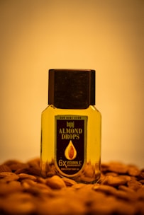 A bottle of Bajaj Almond Drops hair oil is placed on a surface scattered with almonds. The bottle is transparent with a black cap, showing the golden-colored oil inside. The label on the bottle emphasizes '6X Vitamin E' nourishment.