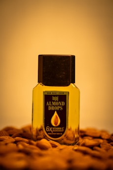 A bottle of Bajaj Almond Drops hair oil is placed on a surface scattered with almonds. The bottle is transparent with a black cap, showing the golden-colored oil inside. The label on the bottle emphasizes '6X Vitamin E' nourishment.