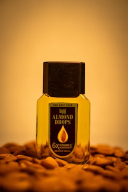 A bottle of Bajaj Almond Drops hair oil is placed on a surface scattered with almonds. The bottle is transparent with a black cap, showing the golden-colored oil inside. The label on the bottle emphasizes '6X Vitamin E' nourishment.