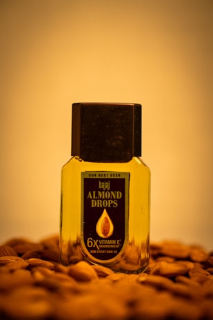 A bottle of Bajaj Almond Drops hair oil is placed on a surface scattered with almonds. The bottle is transparent with a black cap, showing the golden-colored oil inside. The label on the bottle emphasizes '6X Vitamin E' nourishment.