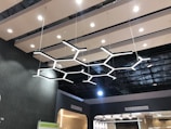A modern ceiling with a striking geometric light fixture made up of interconnected hexagonal shapes. The ceiling consists of parallel wooden slats with recessed lighting. Signs on the walls indicate areas labeled 'Magnetic Lights' and 'Office Automation'.