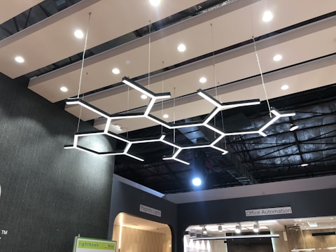 A modern ceiling with a striking geometric light fixture made up of interconnected hexagonal shapes. The ceiling consists of parallel wooden slats with recessed lighting. Signs on the walls indicate areas labeled 'Magnetic Lights' and 'Office Automation'.