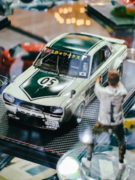 A model car displayed on a reflective surface, featuring a retro design with a dark green and white color scheme. The car has racing decals and a number 05 on the hood. Next to the car is a blurred figure of a person in casual attire, suggesting a dynamic action pose.