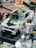A model car displayed on a reflective surface, featuring a retro design with a dark green and white color scheme. The car has racing decals and a number 05 on the hood. Next to the car is a blurred figure of a person in casual attire, suggesting a dynamic action pose.