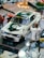 A model car displayed on a reflective surface, featuring a retro design with a dark green and white color scheme. The car has racing decals and a number 05 on the hood. Next to the car is a blurred figure of a person in casual attire, suggesting a dynamic action pose.
