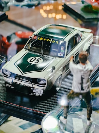 A model car displayed on a reflective surface, featuring a retro design with a dark green and white color scheme. The car has racing decals and a number 05 on the hood. Next to the car is a blurred figure of a person in casual attire, suggesting a dynamic action pose.