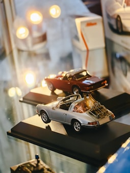 Two miniature car models are displayed on black bases, surrounded by ambient lighting. The cars are highly detailed, reflecting a sense of precision and craft. The scene includes reflections on a glossy surface, contributing to the overall aesthetic.