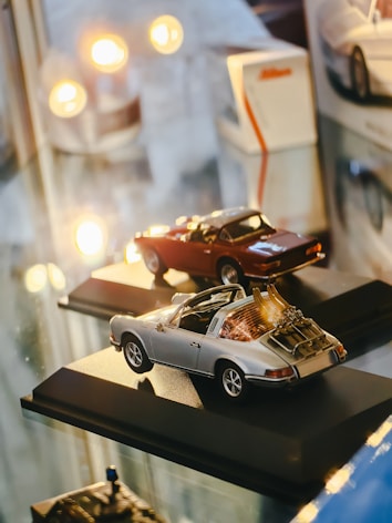 Two miniature car models are displayed on black bases, surrounded by ambient lighting. The cars are highly detailed, reflecting a sense of precision and craft. The scene includes reflections on a glossy surface, contributing to the overall aesthetic.