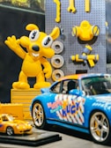 A brightly colored display featuring a yellow cartoon character and various toy cars. The character has large eyes and is raising one arm, surrounded by some toy tools attached to a pegboard. In the foreground, there are two model cars, one larger blue sports car with colorful graphics and a smaller yellow car.