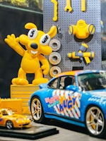 A brightly colored display featuring a yellow cartoon character and various toy cars. The character has large eyes and is raising one arm, surrounded by some toy tools attached to a pegboard. In the foreground, there are two model cars, one larger blue sports car with colorful graphics and a smaller yellow car.