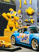 A brightly colored display featuring a yellow cartoon character and various toy cars. The character has large eyes and is raising one arm, surrounded by some toy tools attached to a pegboard. In the foreground, there are two model cars, one larger blue sports car with colorful graphics and a smaller yellow car.
