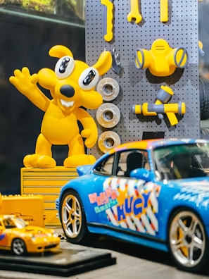 A brightly colored display featuring a yellow cartoon character and various toy cars. The character has large eyes and is raising one arm, surrounded by some toy tools attached to a pegboard. In the foreground, there are two model cars, one larger blue sports car with colorful graphics and a smaller yellow car.
