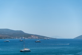 A tranquil ocean scene with several boats floating on clear blue water. Green hills line the distant horizon under a bright blue sky. A catamaran is prominently visible in the foreground.
