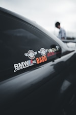 A close-up of a car window featuring various stickers including the logos 'BMW NL Enthusiasts' and 'BADD'. The reflection on the window shows a blurred image of a person standing nearby.
