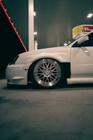 A white car with modified features is parked at a gas station. The focus is on the car’s large, shiny alloy wheel and low-profile tire. The scene is illuminated by the artificial lighting of the gas station, casting soft shadows on the concrete ground. In the background, there is a Shell gas station sign.
