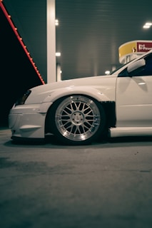 A white car with modified features is parked at a gas station. The focus is on the car’s large, shiny alloy wheel and low-profile tire. The scene is illuminated by the artificial lighting of the gas station, casting soft shadows on the concrete ground. In the background, there is a Shell gas station sign.