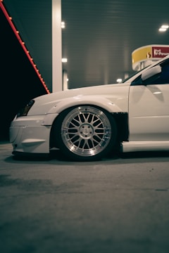 A white car with modified features is parked at a gas station. The focus is on the car’s large, shiny alloy wheel and low-profile tire. The scene is illuminated by the artificial lighting of the gas station, casting soft shadows on the concrete ground. In the background, there is a Shell gas station sign.