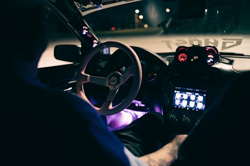 A nighttime view of a car interior with ambient purple LED lighting. The dashboard displays digital gauges with red and blue lighting alongside a touchscreen interface. A partially visible person sits in the driver's seat, hands on the steering wheel. Outside lights faintly illuminate the road ahead.