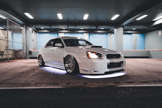A well-modified white car is parked in a dimly lit garage, with distinctive alloy wheels and underglow lighting. The environment features a gritty, urban industrial style with chain-link fencing and concrete floors.
