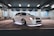 A well-modified white car is parked in a dimly lit garage, with distinctive alloy wheels and underglow lighting. The environment features a gritty, urban industrial style with chain-link fencing and concrete floors.