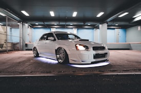 A well-modified white car is parked in a dimly lit garage, with distinctive alloy wheels and underglow lighting. The environment features a gritty, urban industrial style with chain-link fencing and concrete floors.