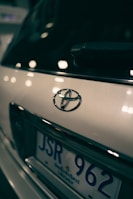 Close-up view of a car's rear, focusing on the Toyota emblem and part of the license plate with a blurred background. The lighting creates reflections and soft highlights on the vehicle's surface.