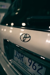 Close-up view of a car's rear, focusing on the Toyota emblem and part of the license plate with a blurred background. The lighting creates reflections and soft highlights on the vehicle's surface.