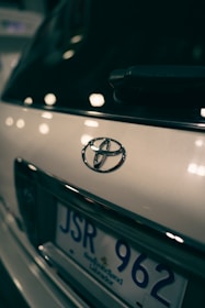 Close-up view of a car's rear, focusing on the Toyota emblem and part of the license plate with a blurred background. The lighting creates reflections and soft highlights on the vehicle's surface.