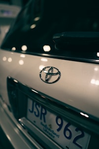 Close-up view of a car's rear, focusing on the Toyota emblem and part of the license plate with a blurred background. The lighting creates reflections and soft highlights on the vehicle's surface.