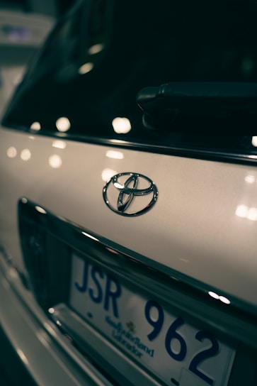 Close-up view of a car's rear, focusing on the Toyota emblem and part of the license plate with a blurred background. The lighting creates reflections and soft highlights on the vehicle's surface.