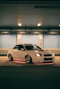 A white car with custom modifications is parked in a dimly lit urban setting. The vehicle features underglow neon lights that emit a red hue, highlighting its sporty design and aftermarket wheels. The scene has a soft blue and orange ambience due to overhead lighting.