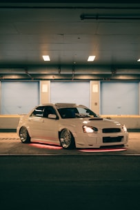 A white car with custom modifications is parked in a dimly lit urban setting. The vehicle features underglow neon lights that emit a red hue, highlighting its sporty design and aftermarket wheels. The scene has a soft blue and orange ambience due to overhead lighting.