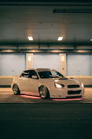 A white car with custom modifications is parked in a dimly lit urban setting. The vehicle features underglow neon lights that emit a red hue, highlighting its sporty design and aftermarket wheels. The scene has a soft blue and orange ambience due to overhead lighting.