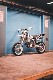 A detailed view of a white and black dirt bike positioned against a light blue garage door in an industrial setting. The bike features racing decals and is propped on a concrete floor. Overhead, exposed pipes run along the ceiling, and two warning signs are posted on the surrounding walls.