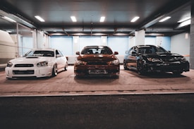 Three modified sports cars are parked side by side in an indoor garage with industrial lighting. The left car is white, the middle car has a brownish-red color, and the right car is black. The setting has a gritty urban feel with concrete floors and chain-link fencing along the walls.