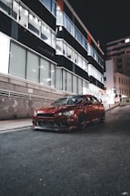 A sleek black sports car parked under neon red streetlights at night.