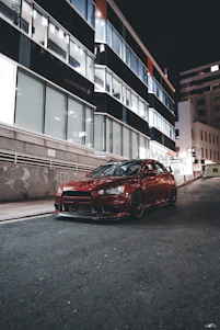 A sleek crimson luxury car parked on a city street at dusk, with soft urban lights reflecting off its polished surface.