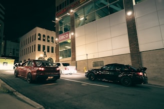 Two cars are parked on a dimly lit urban street at night. The surrounding buildings include a bank with prominent signage and a gym with illuminated letters. The atmosphere is quiet, and the empty street is lit by streetlights and the glow from the buildings.