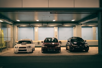 Three cars are parked closely side by side in an underground parking area. The setting is dimly lit with overhead lights illuminating the vehicles. The background features a blue industrial-style wall with large closed doors.