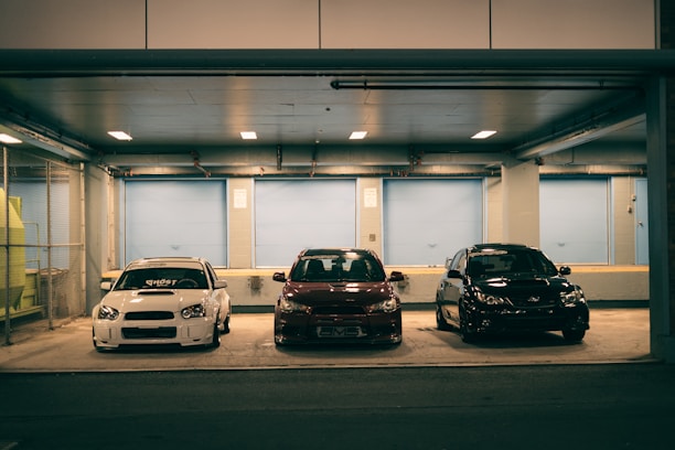 Three cars are parked closely side by side in an underground parking area. The setting is dimly lit with overhead lights illuminating the vehicles. The background features a blue industrial-style wall with large closed doors.