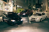 A lineup of modified JDM cars parked at an urban car meet under neon streetlights.