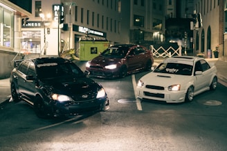 Three modified cars are parked on an urban street at night. The surroundings include buildings, neon lights, and a visible street sign. The scene is dimly lit, highlighting the cars' features.