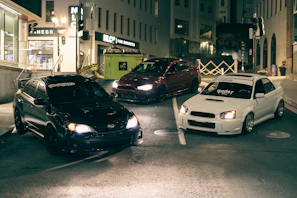 A group of modified JDM cars lined up at a nighttime car meet with neon reflections.