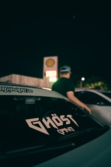 A car's rear window displays a sticker with the text 'Ghost Gang' in stylized font. A person wearing a cap stands near the car, with the background showing a Shell gas station sign illuminated against the night sky.