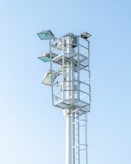 A tall metal structure with several mounted floodlights, standing against a clear blue sky. The structure has a ladder for maintenance access and is surrounded by protective railings.