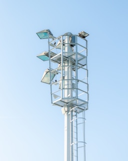 A tall metal structure with several mounted floodlights, standing against a clear blue sky. The structure has a ladder for maintenance access and is surrounded by protective railings.