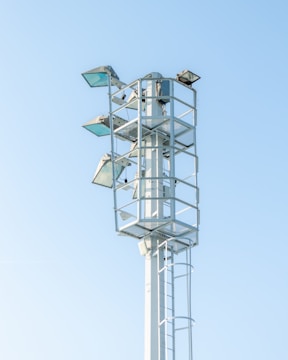 A tall metal structure with several mounted floodlights, standing against a clear blue sky. The structure has a ladder for maintenance access and is surrounded by protective railings.
