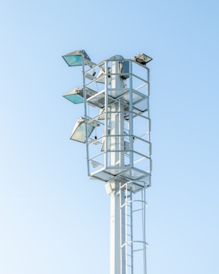 A tall metal structure with several mounted floodlights, standing against a clear blue sky. The structure has a ladder for maintenance access and is surrounded by protective railings.