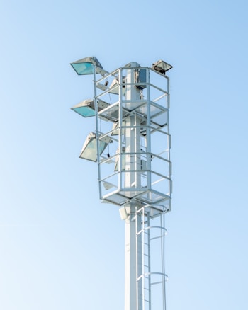 A tall metal structure with several mounted floodlights, standing against a clear blue sky. The structure has a ladder for maintenance access and is surrounded by protective railings.
