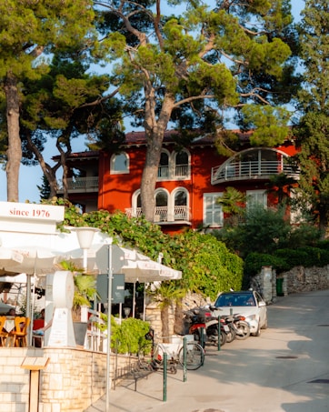 A vibrant orange house with white railings is set against a backdrop of tall pine trees. In the foreground, a cozy outdoor cafe with white umbrellas and wooden chairs is visible. Several motorbikes and a white car are parked nearby along a paved path lined with greenery and stone walls. The atmosphere is tranquil and sunlit, indicating late afternoon or early evening.